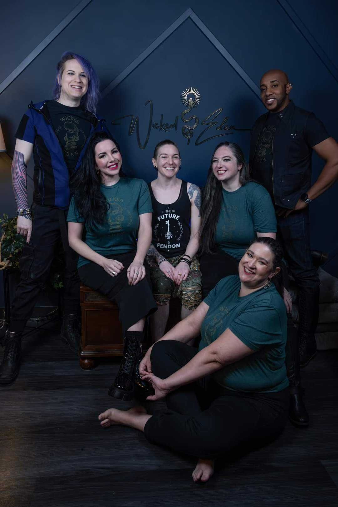 Group of six people posing; dark teal and black clothing; dark blue background.