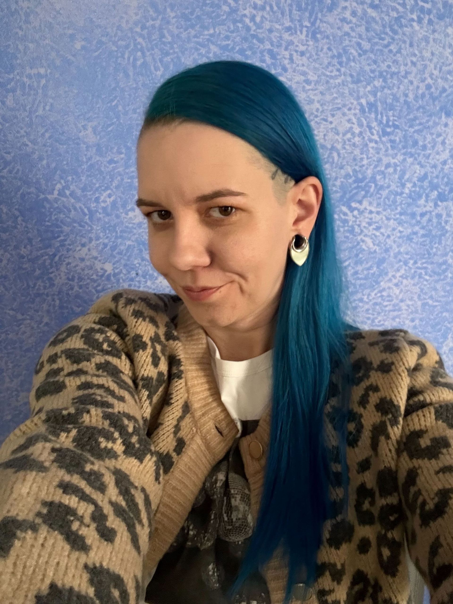 Woman with blue hair in a leopard print sweater, looking at the camera with a slight smile. Blue wall in the background.