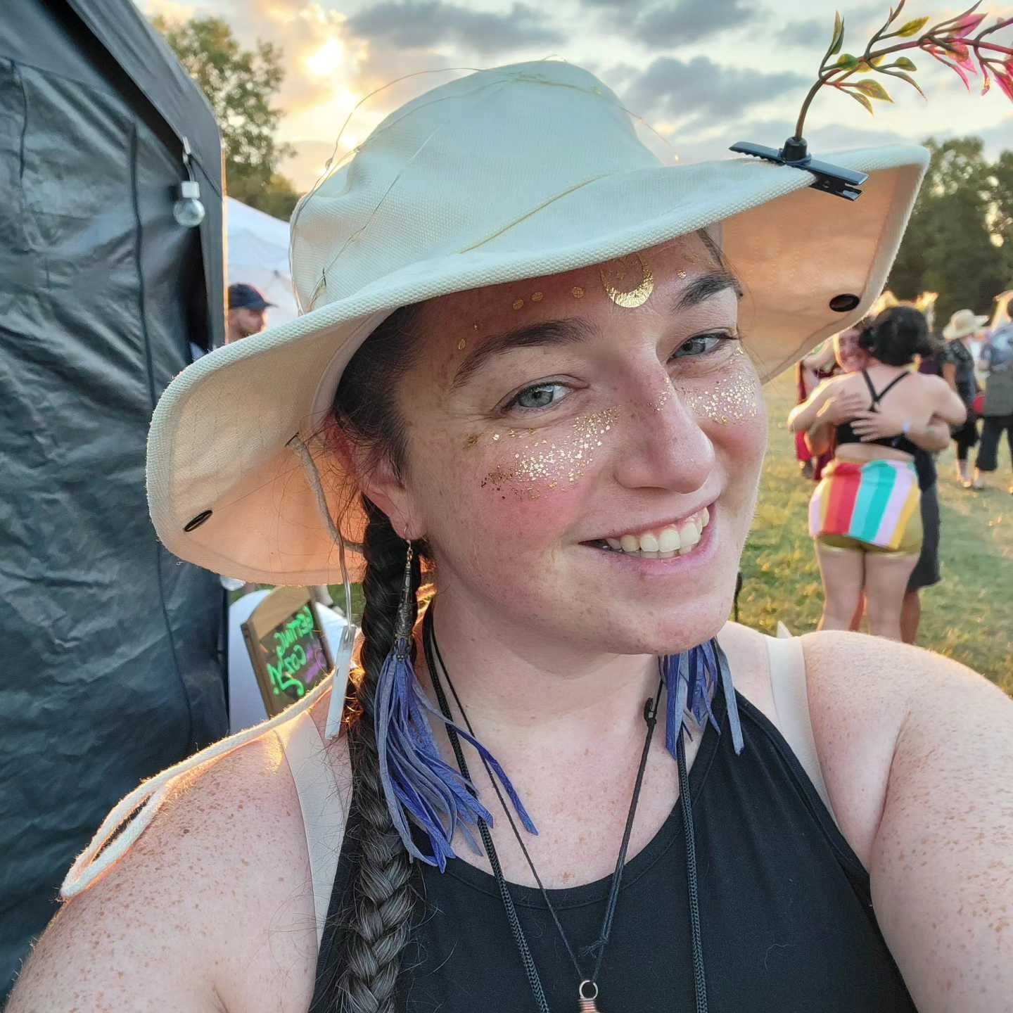 Woman smiling, wearing a hat with floral decoration, glitter makeup, and blue fringe earrings, outside.
