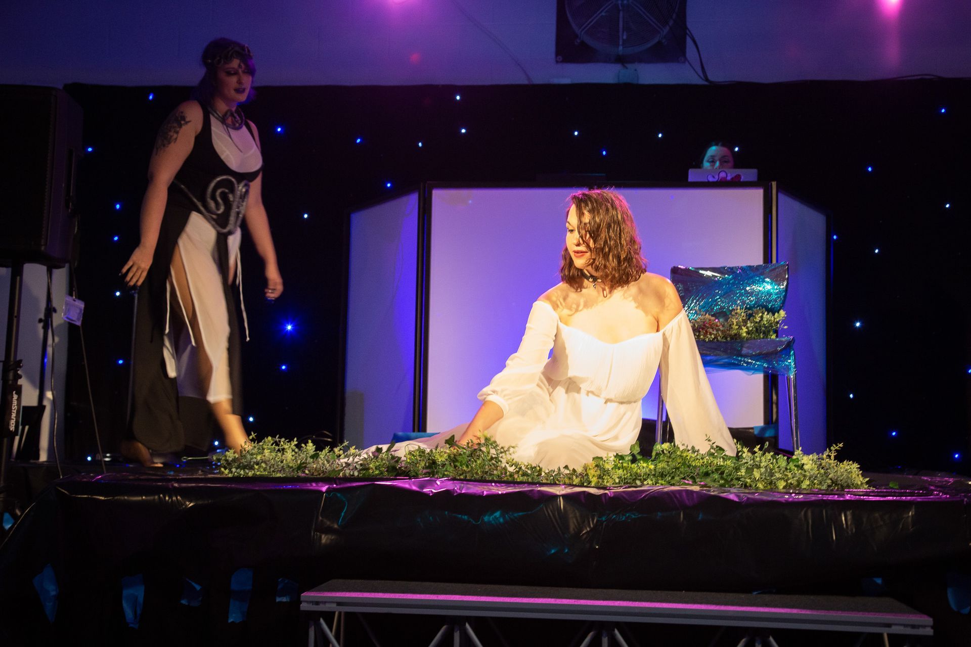 Woman in white gown reclines in foliage on stage; another woman looks on. Black background with lights.