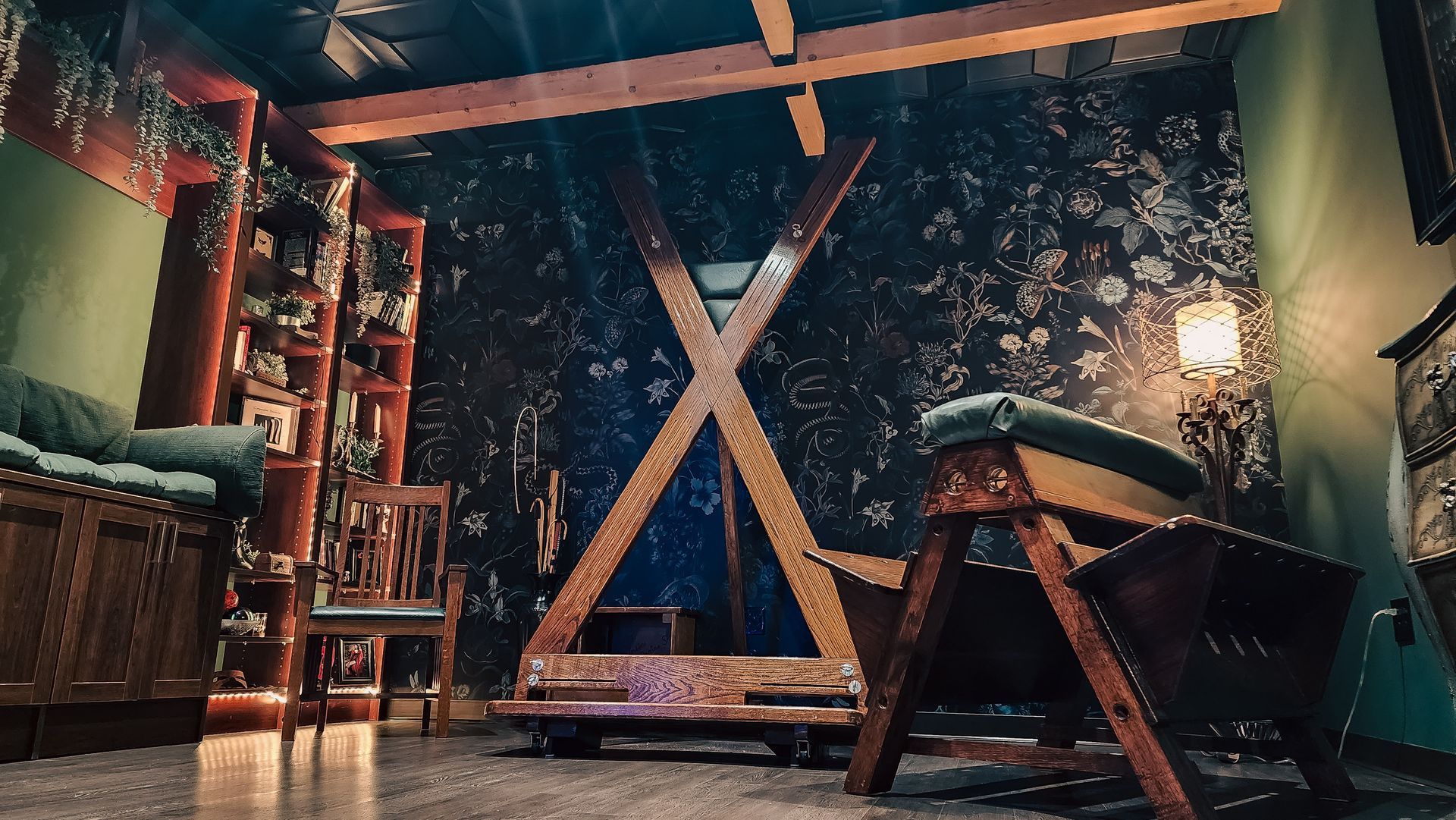 A dimly lit room with BDSM equipment: a cross, bench, and chair, plus dark floral wallpaper.