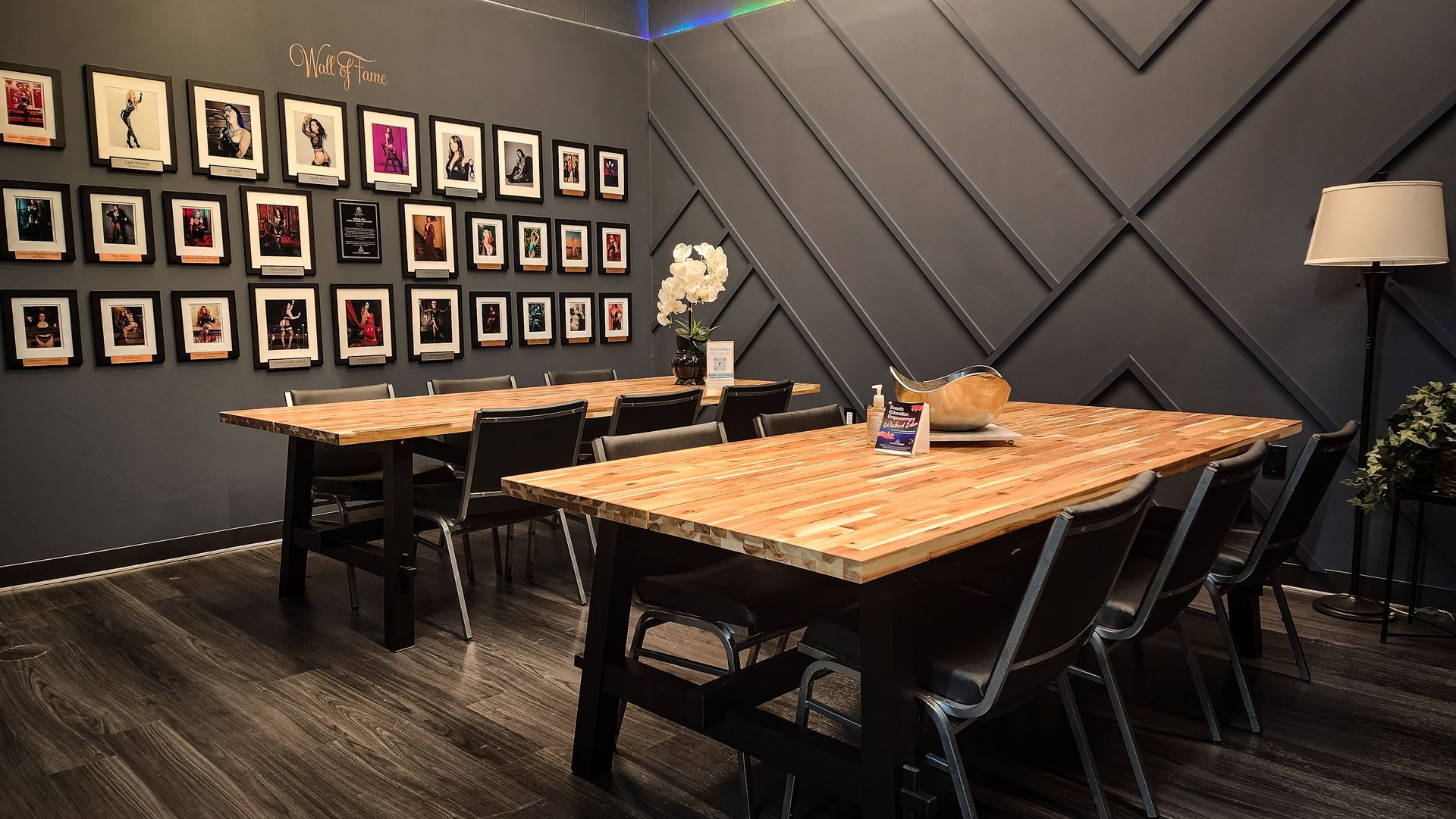 Interior with wooden tables, black chairs, artwork on a dark blue wall. A lamp and greenery add decor.