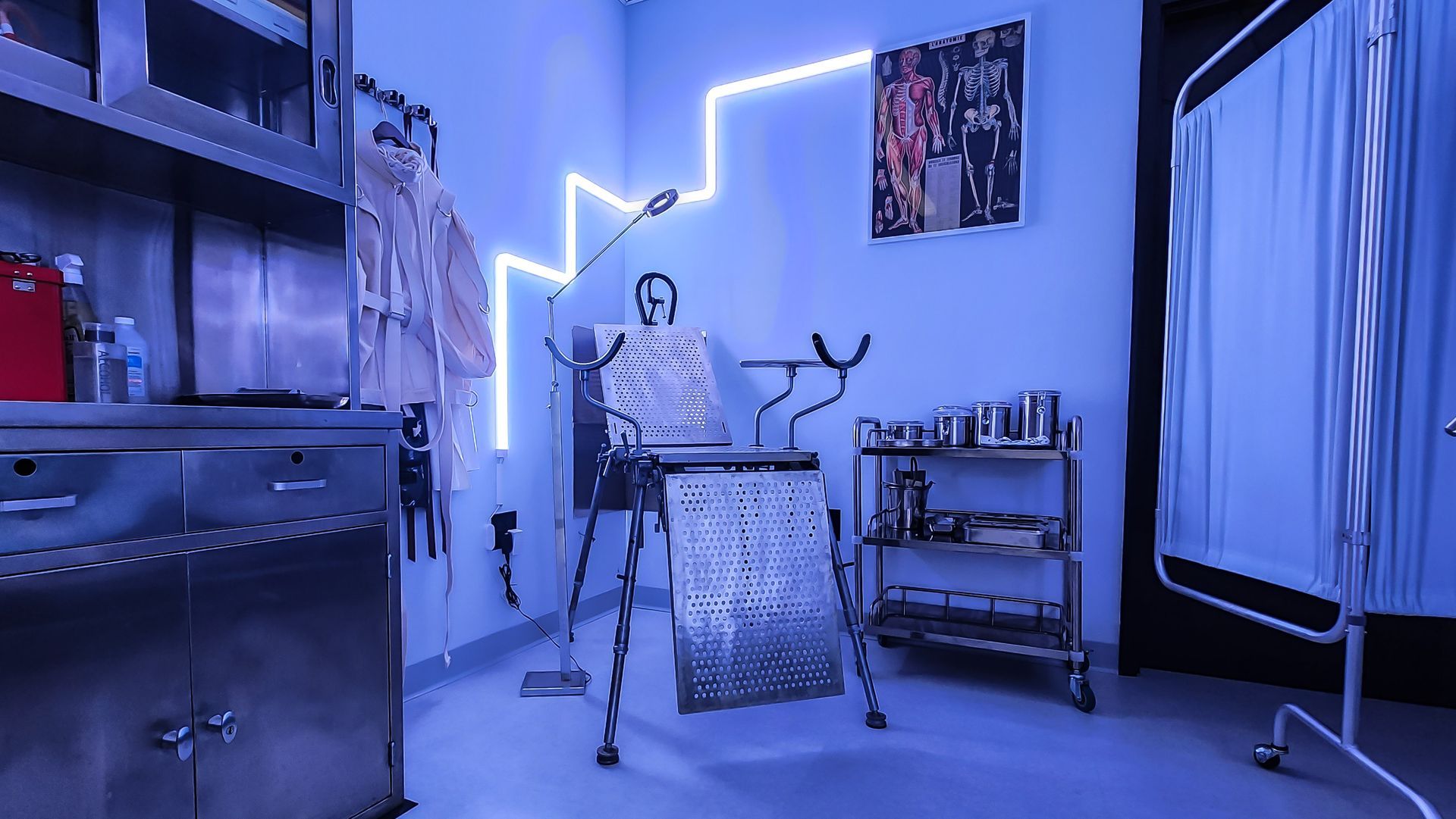 A clinical examination room bathed in blue light; metal exam table, cabinets, and medical supplies.