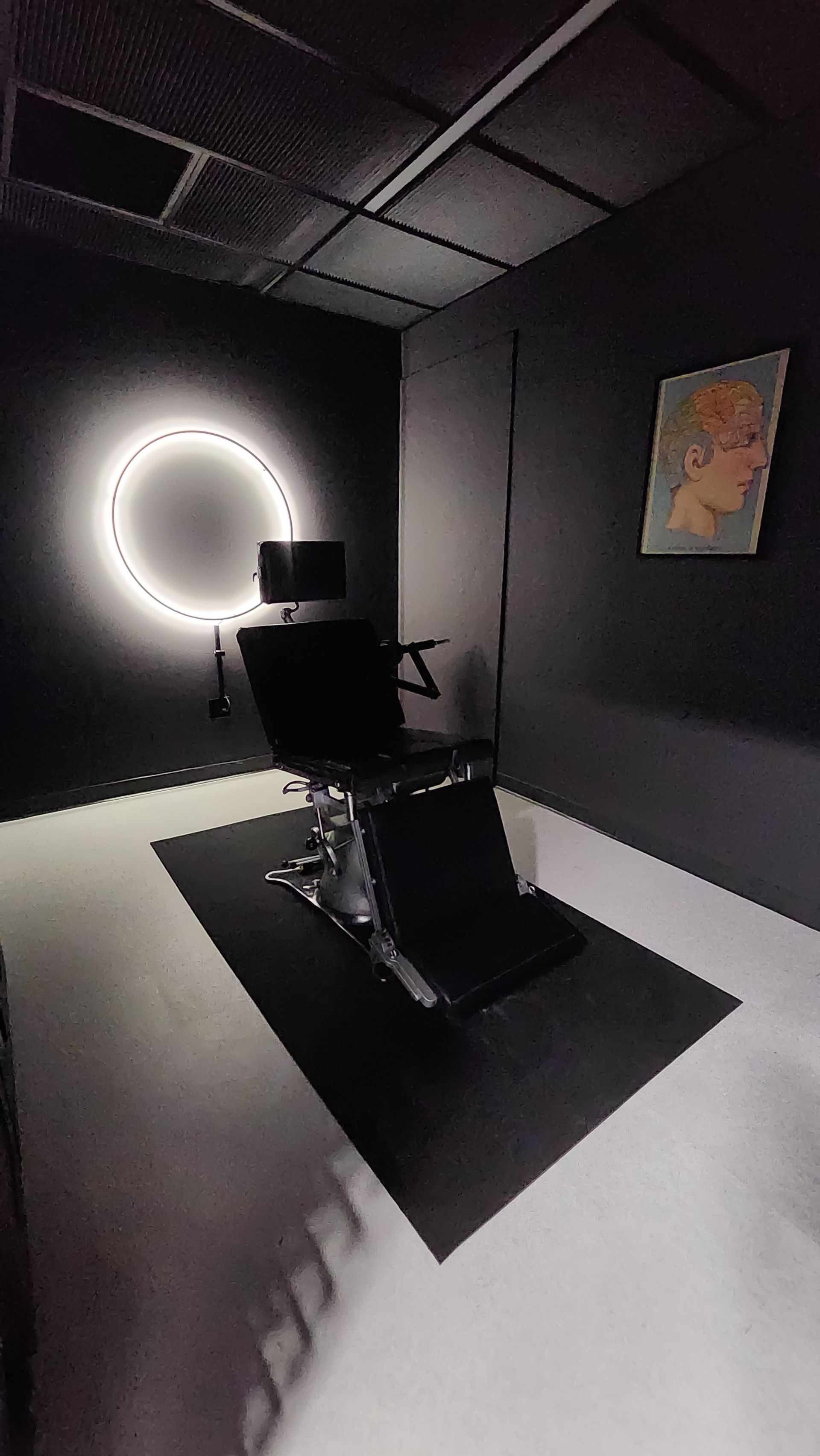 Dark-themed tattoo studio with black chair, a neon circle light, and painting on a black wall.