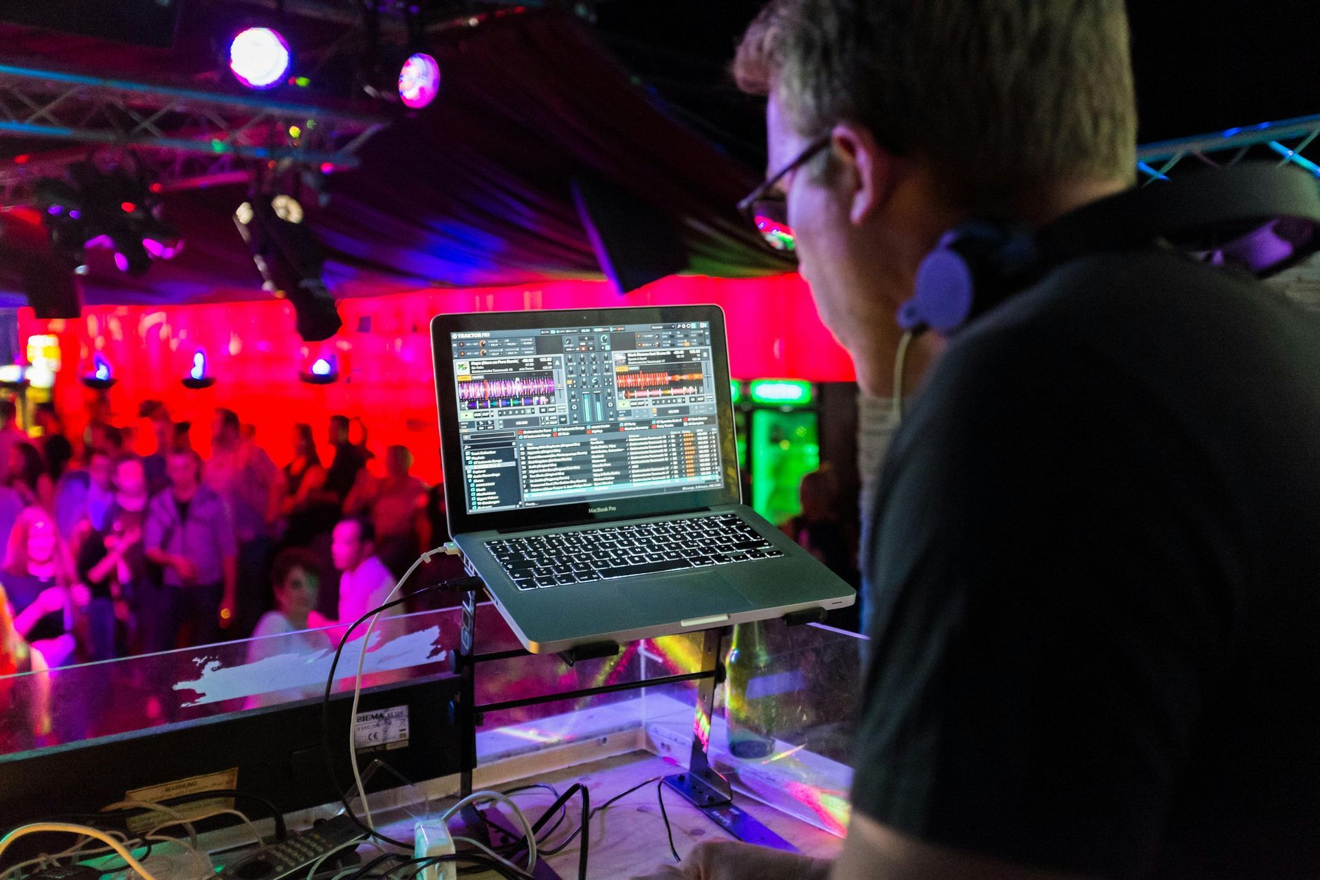 DJ at a club, working on laptop. Red and purple stage lighting, people in the background.