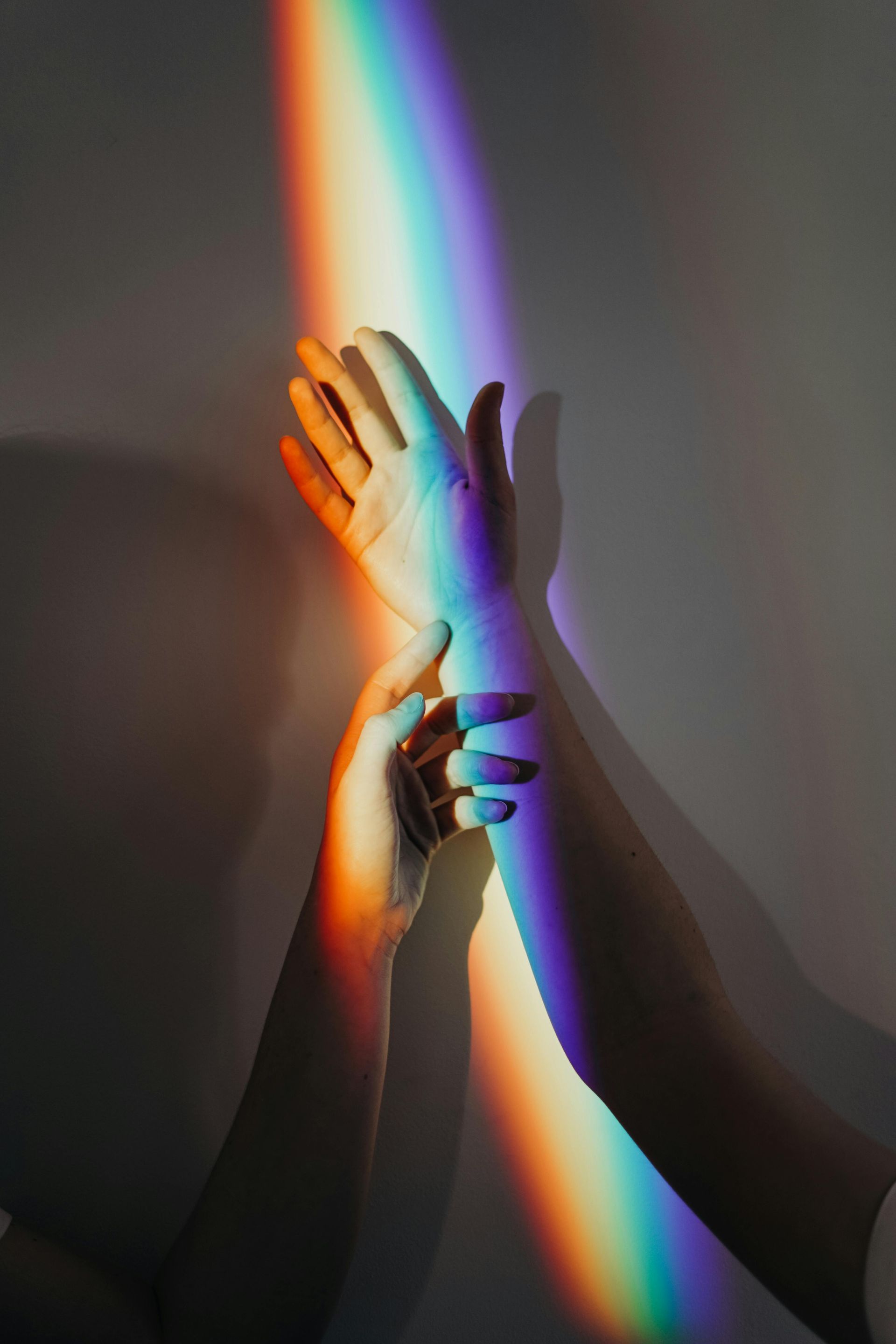 Hands reaching toward a rainbow beam of light on a white wall, creating colorful shadows.