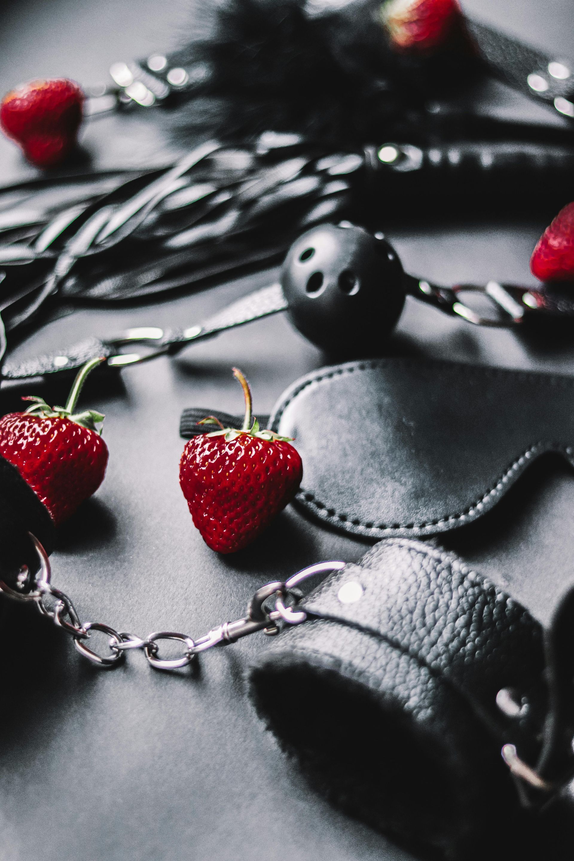 Black leather restraints, blindfold, and whip with strawberries on a dark surface.