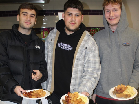 Three people stand in a kitchen, smiling and holding plates of cooked food.