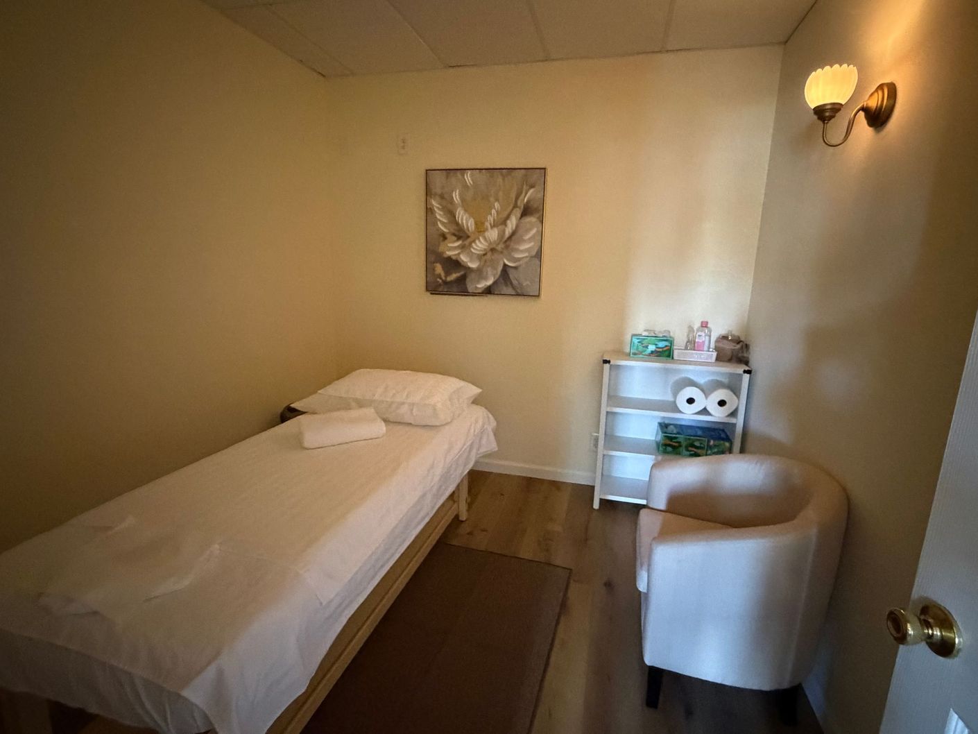 Small treatment room with a single bed, chair, side table, and wall art in soft beige tones