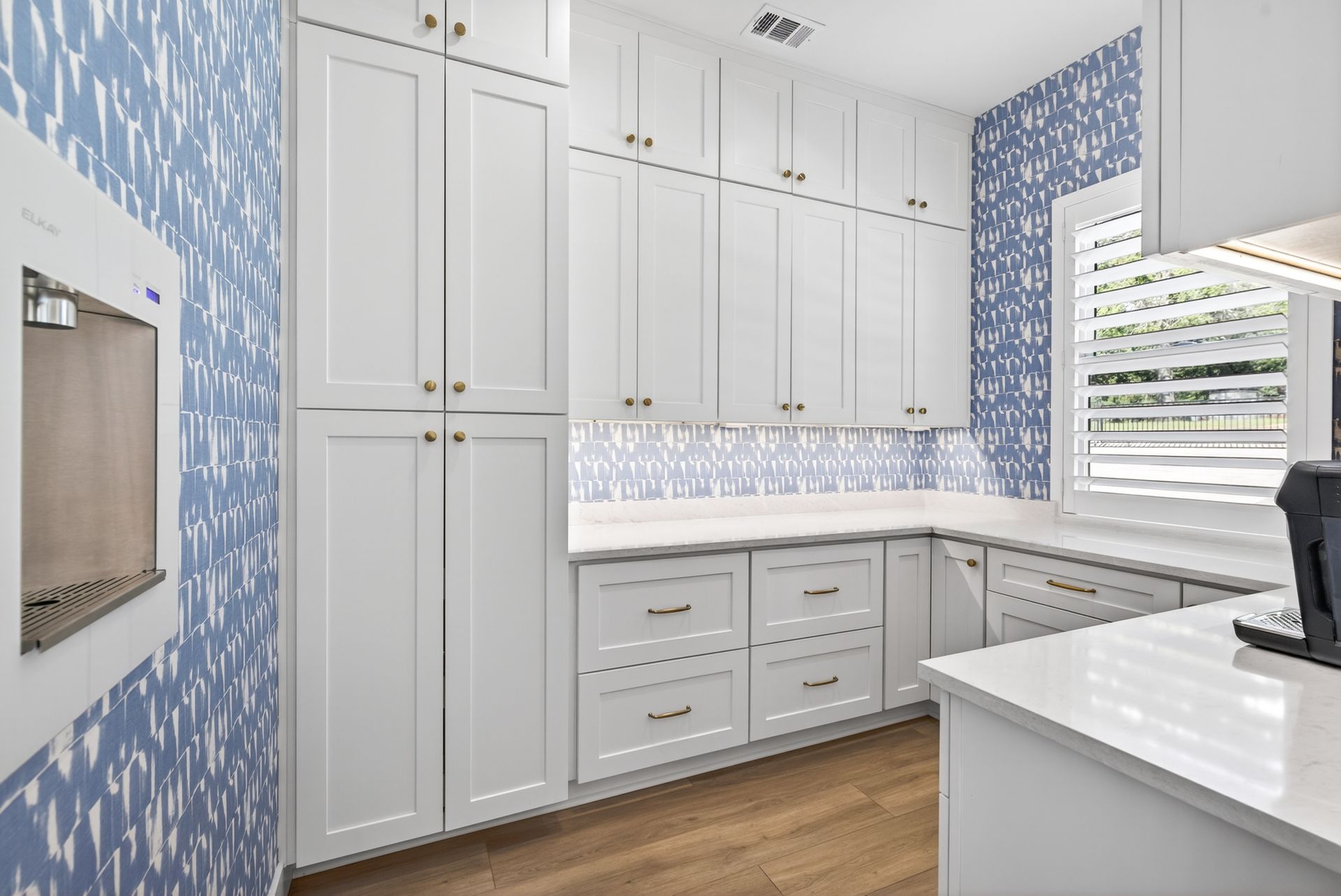 Bright white kitchen with blue patterned wallpaper, white cabinets, wood floor, and a window over the counter