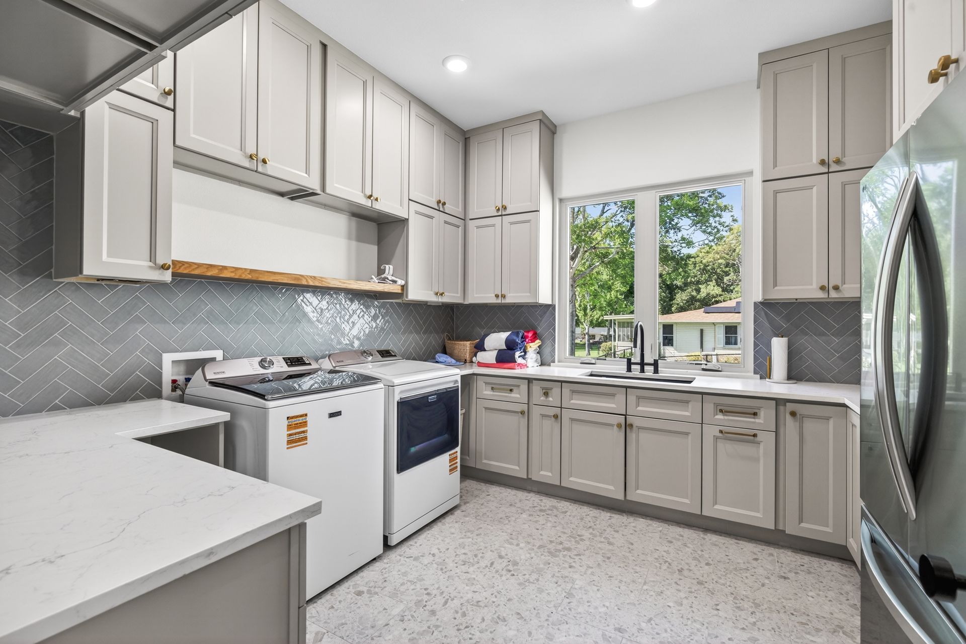 Bright modern kitchen with white cabinets, gray backsplash, stainless fridge, and a window over the sink.