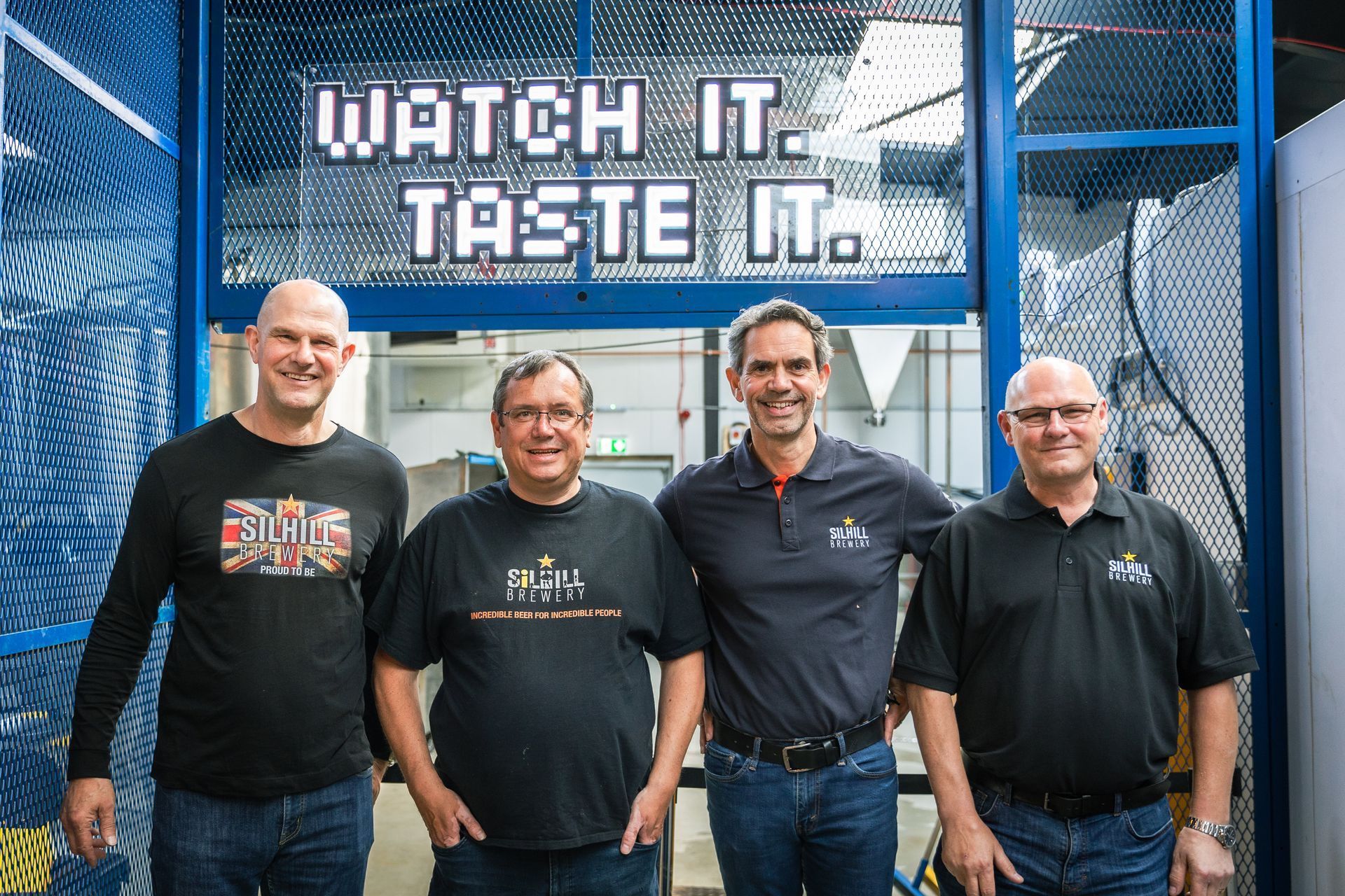 Four men standing in front of a sign that says watch it taste it
