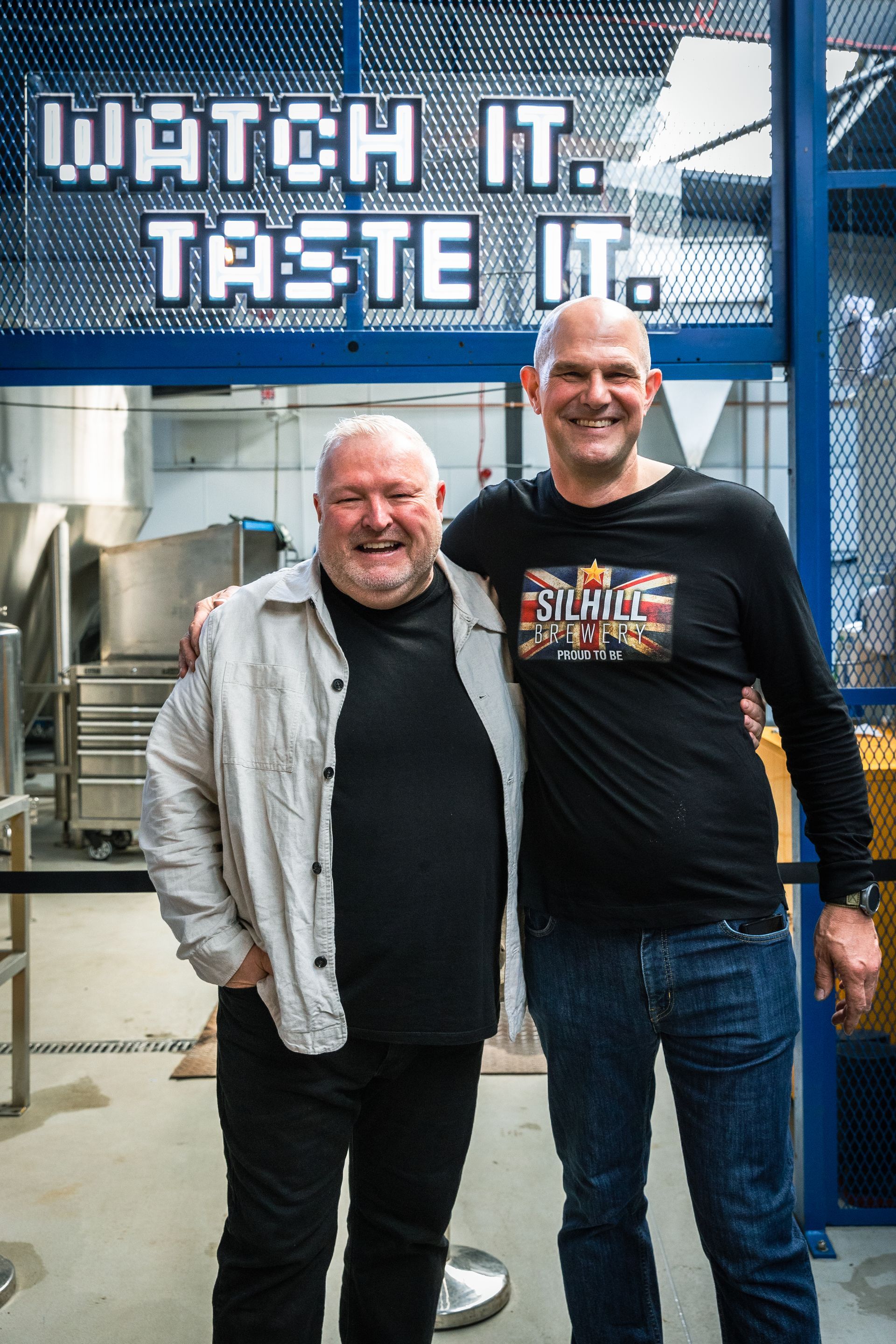 Two men are posing for a picture in front of a sign that says watch it taste it