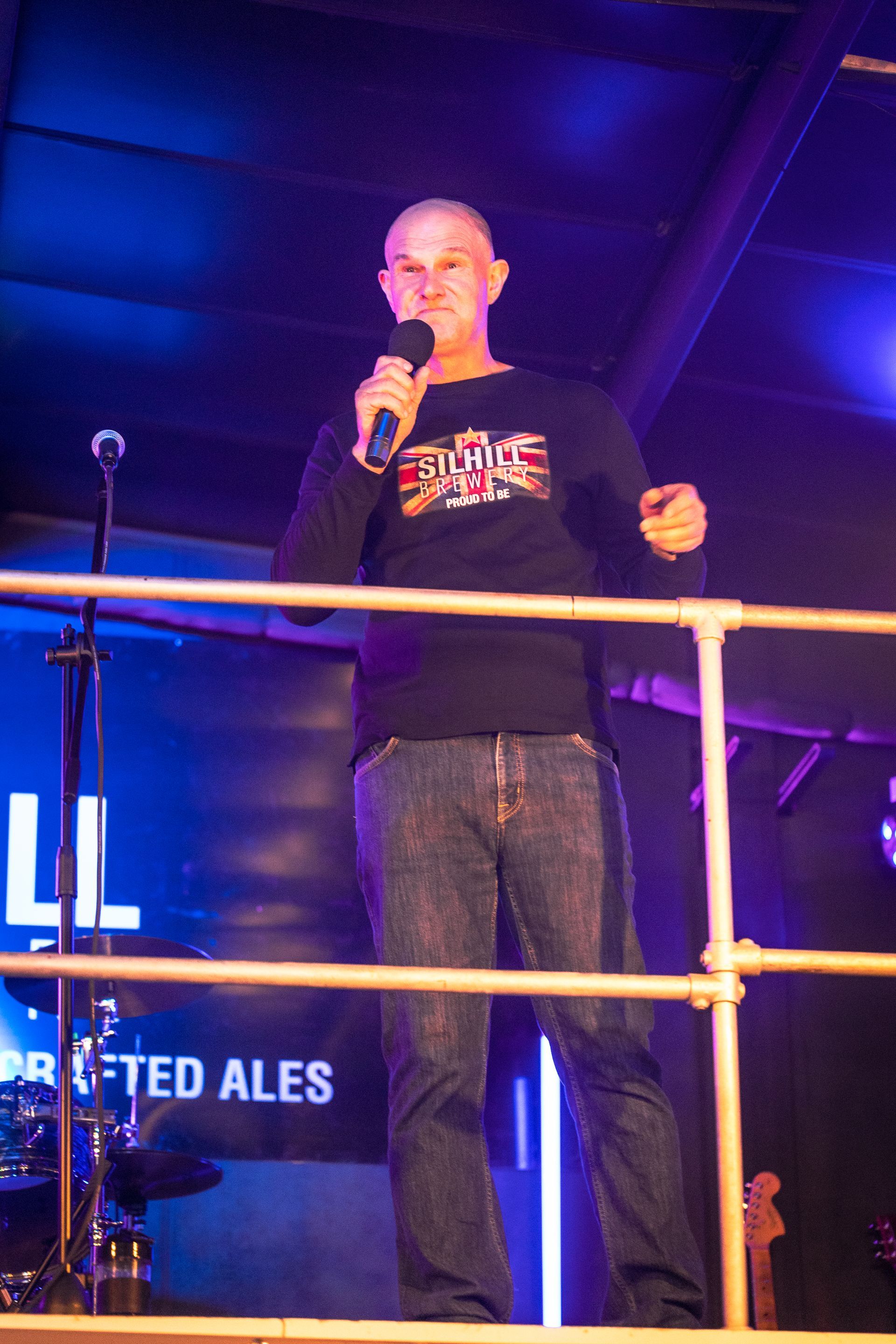 A man stands on a stage holding a microphone in front of a sign that says crafted ales