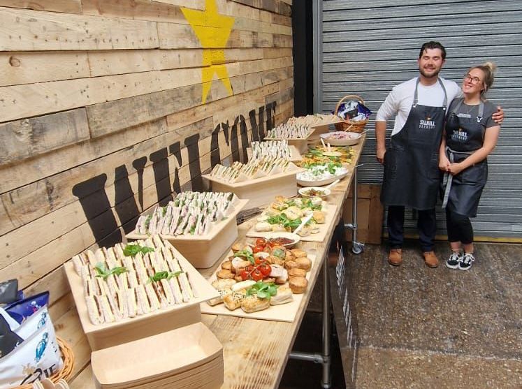 A man and a woman are standing in front of a table with food on it.