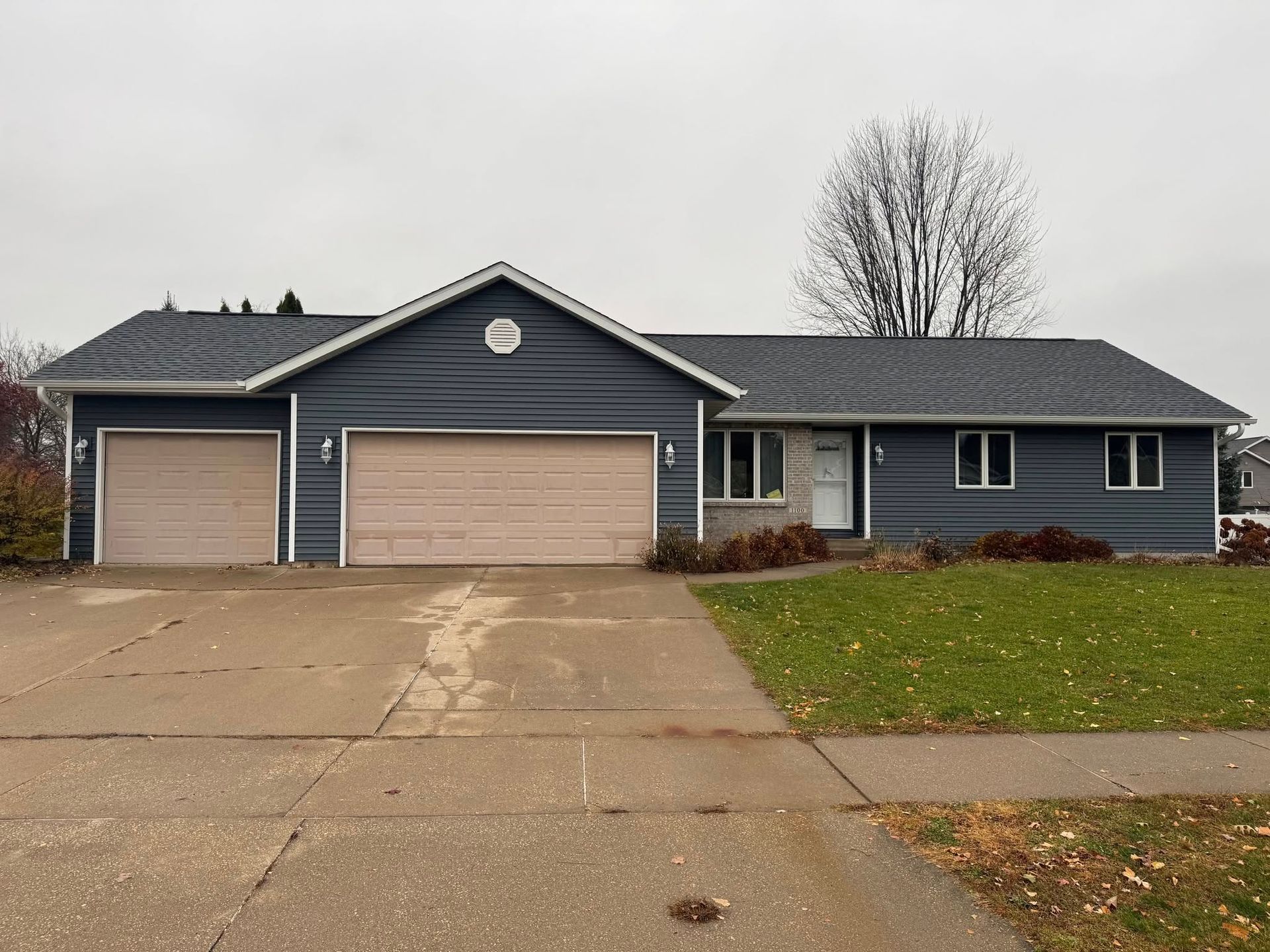 Suburban blue house with two garages, a wide driveway, and a leafless tree in front.