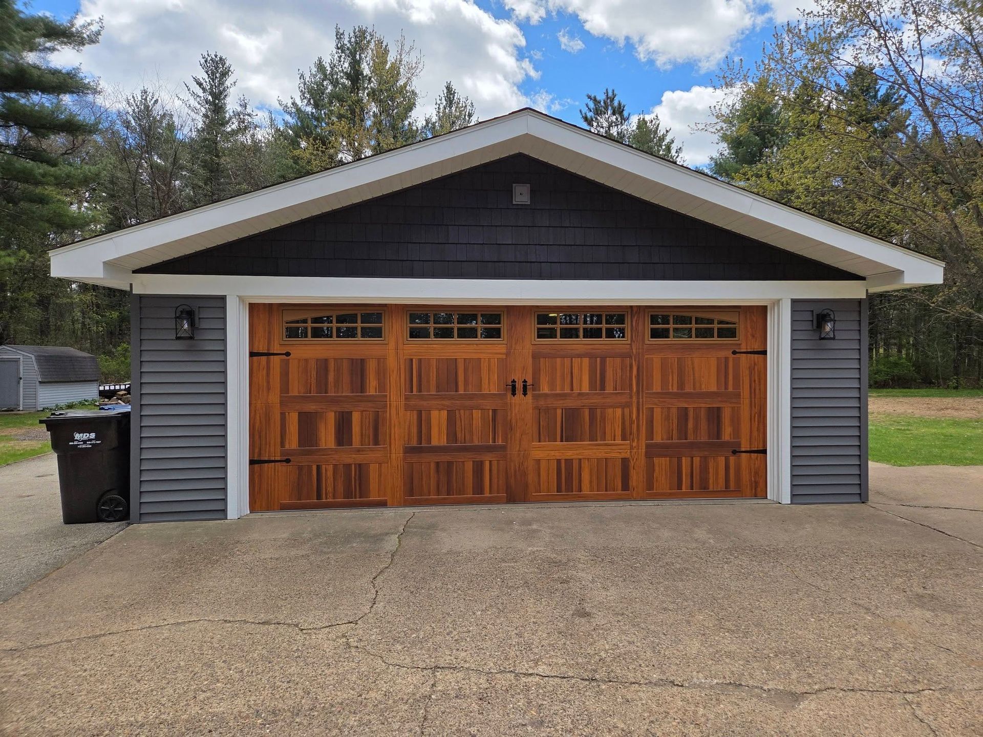 Detached garage with double wooden doors, gray siding, and a black roof surrounded by trees