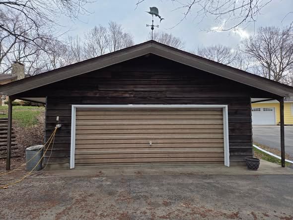 Single-car garage with a dark brown roof and tan door in a driveway, bare trees in the background