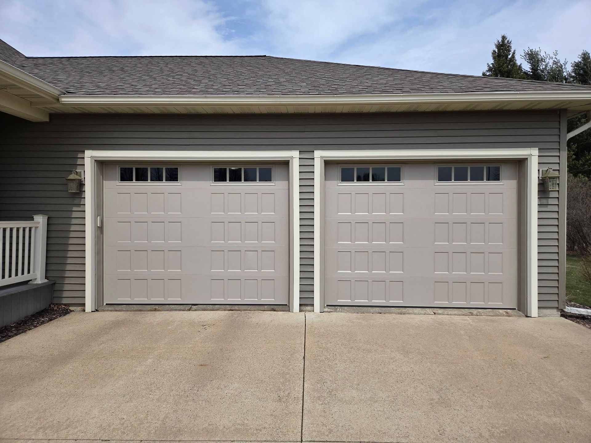 Two gray garage doors with white trim on a house exterior, beside a concrete driveway.