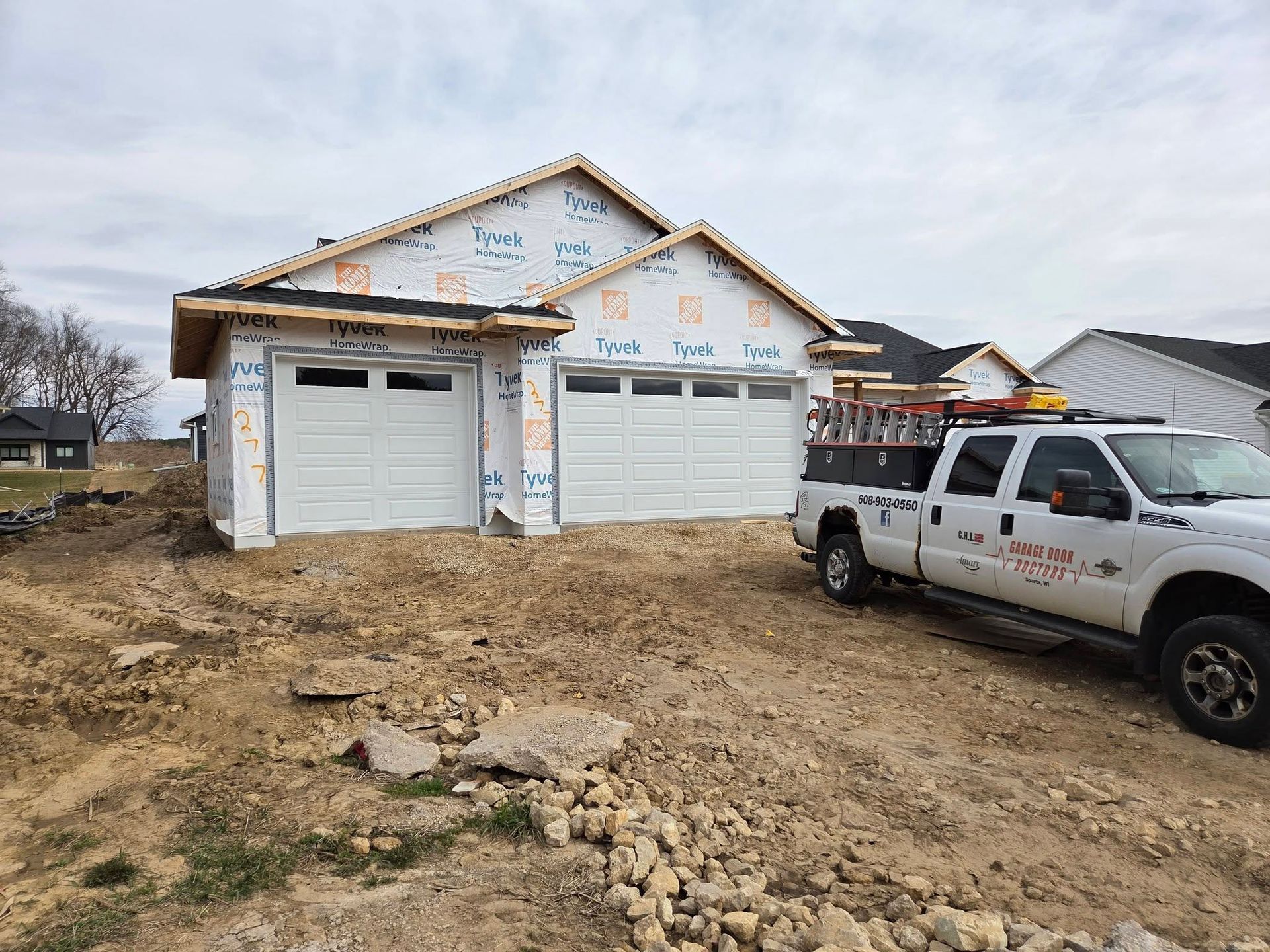 New house under construction with white garage doors and a white pickup truck on a muddy lot