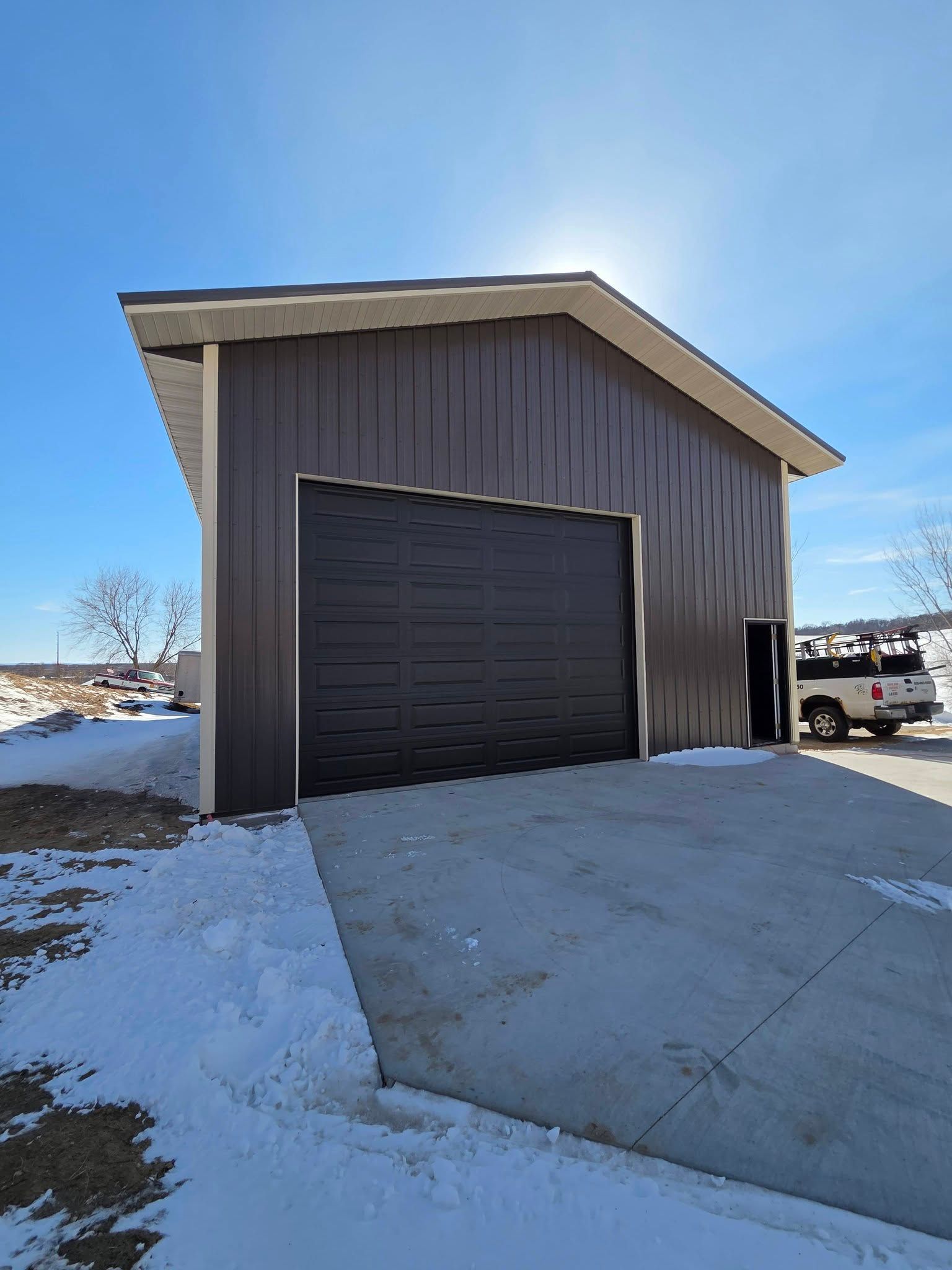Snow-covered metal garage with a black roll-up door and sloped concrete driveway under a clear blue sky.