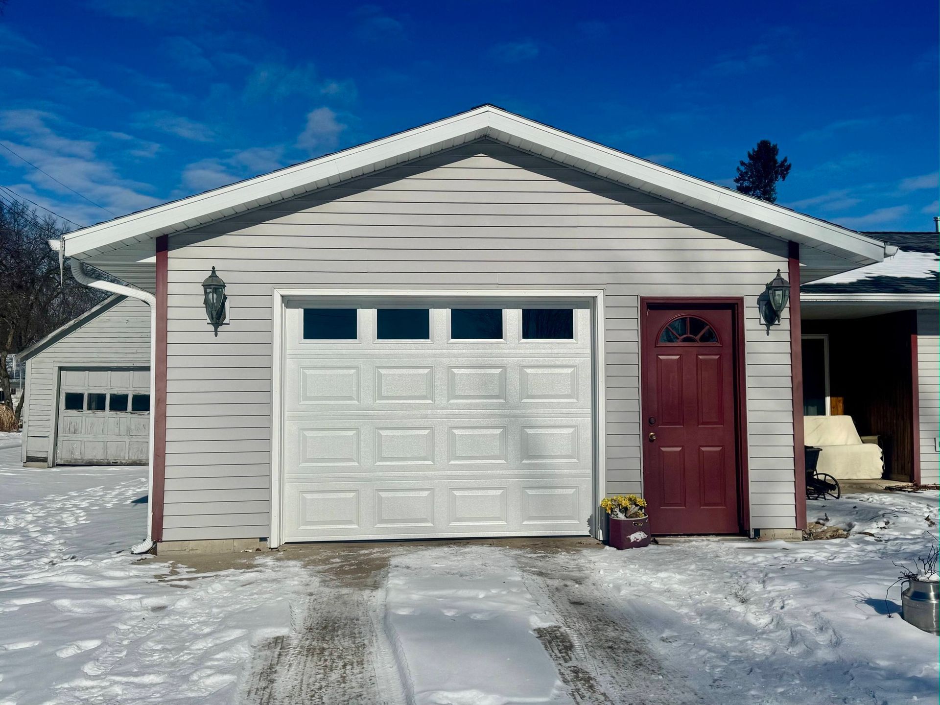 Snowy driveway in front of a beige garage with a white overhead door and red side entrance door
