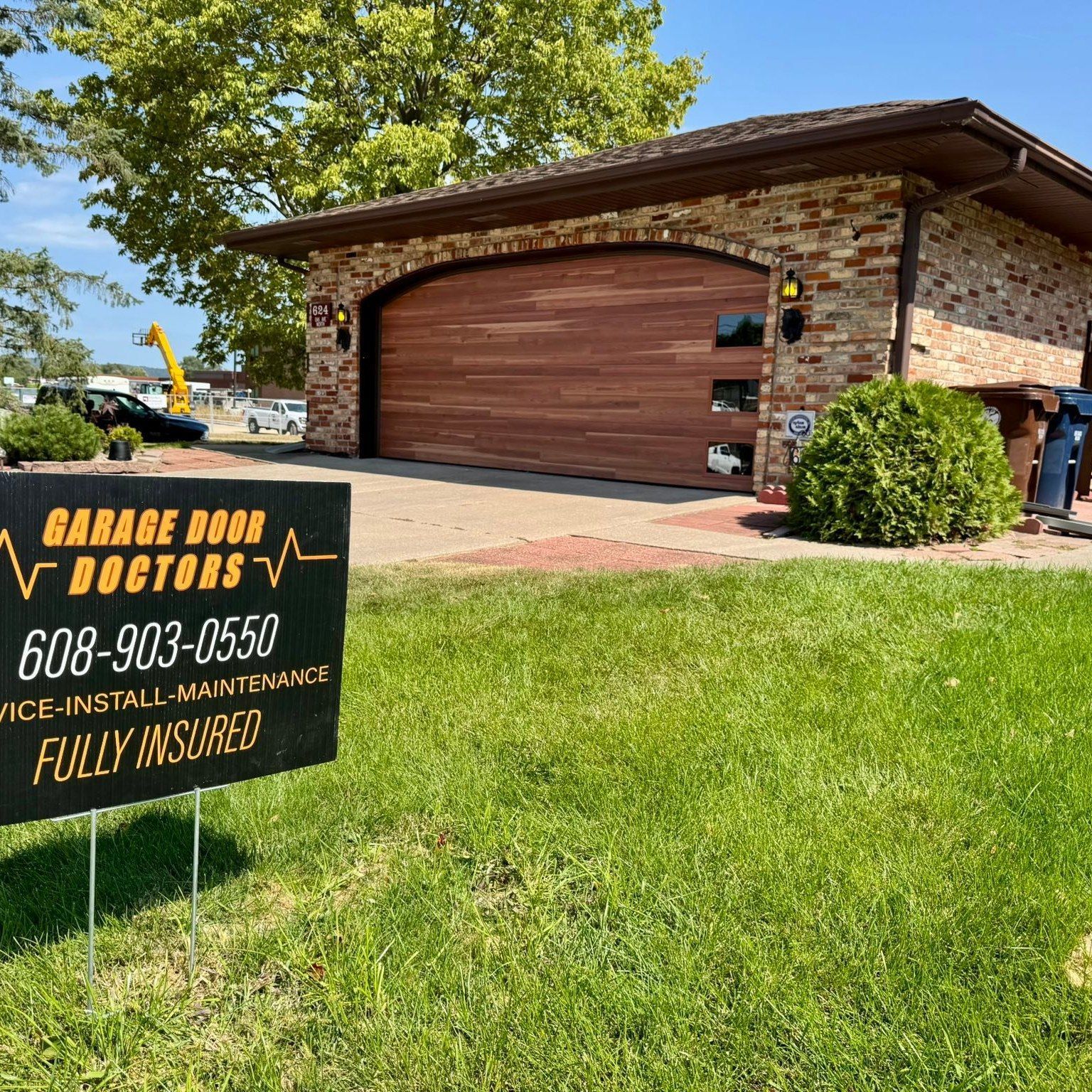 Garage doors doctors sign in front of a brick house with a brown garage door and green lawn.