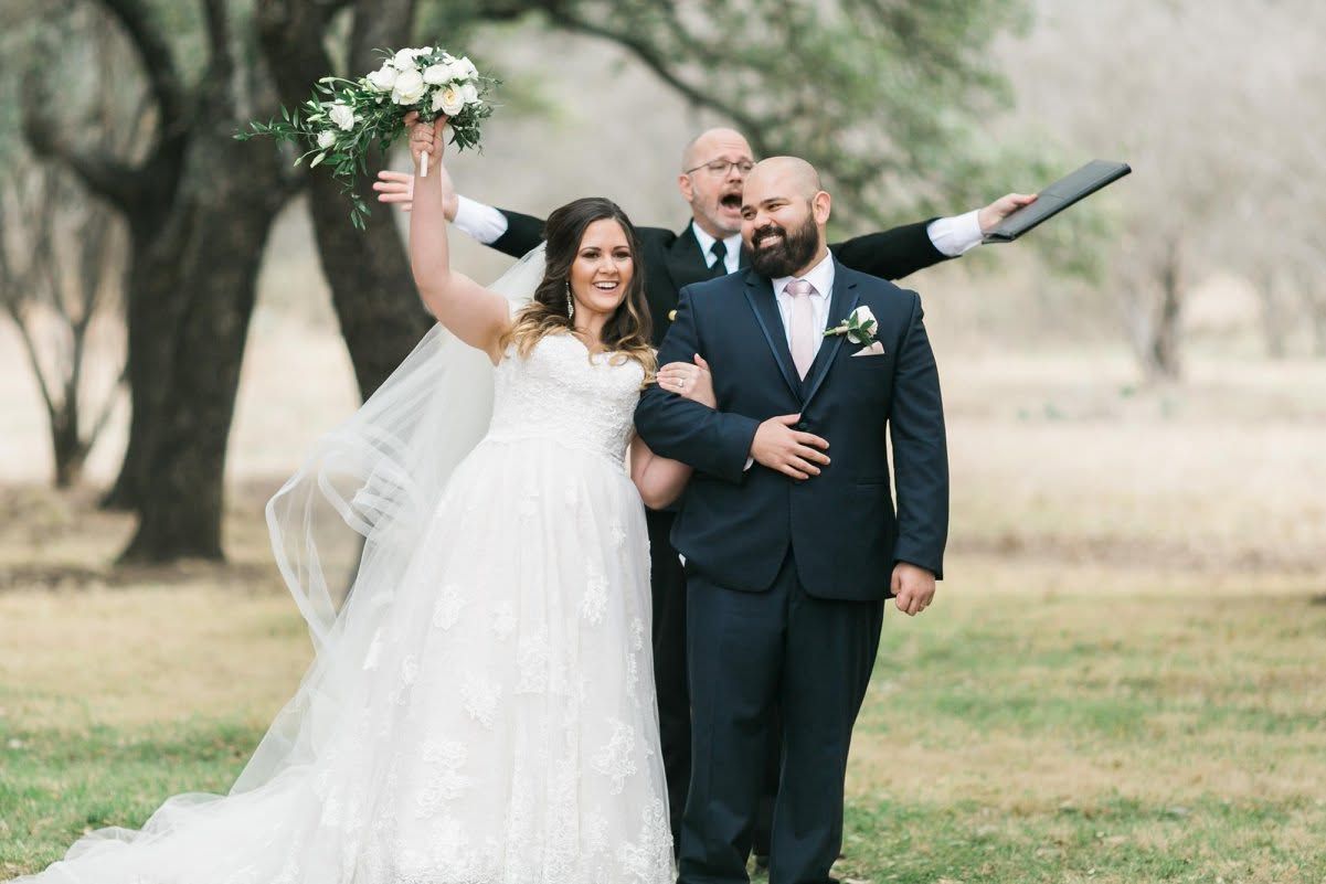 Wedding couple walking outdoors, bride in white dress and groom in navy suit, with a bouquet raised in celebration