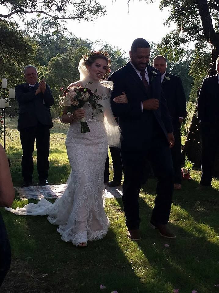 Bride and groom standing outdoors at a wedding ceremony, holding hands in sunlight.