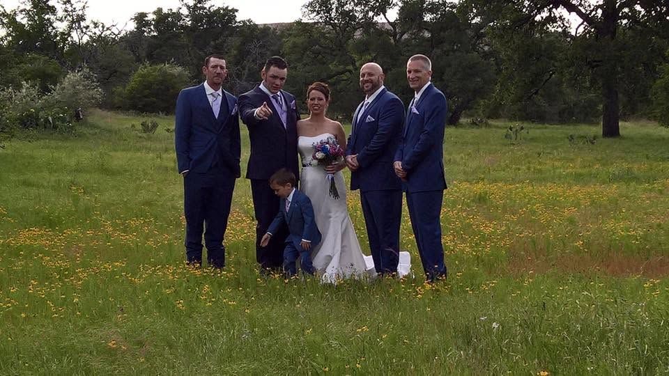 Wedding party posing outdoors in a grassy field, with bride and groom surrounded by four groomsmen.