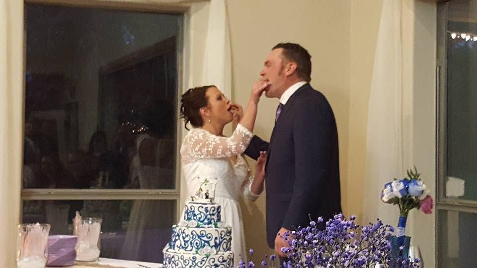 Bride touching groom’s chin beside a decorated wedding cake, smiling indoors