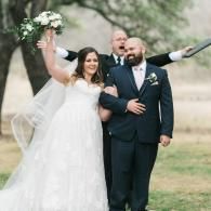 Bride in white dress and groom in navy suit walk outdoors, smiling, with bouquet raised and officiant behind them