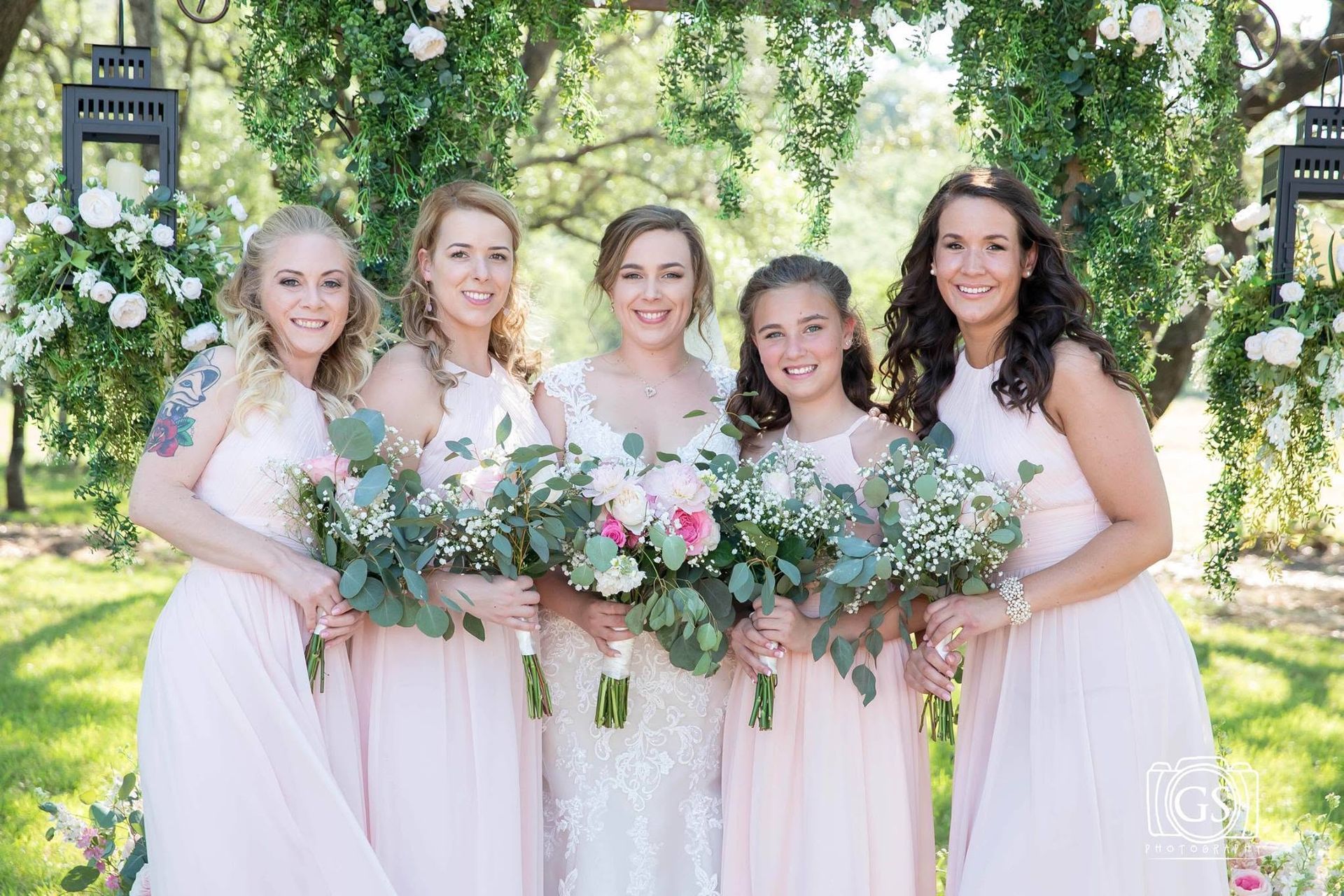 Five women in white dresses holding pastel flower bouquets outdoors under trees