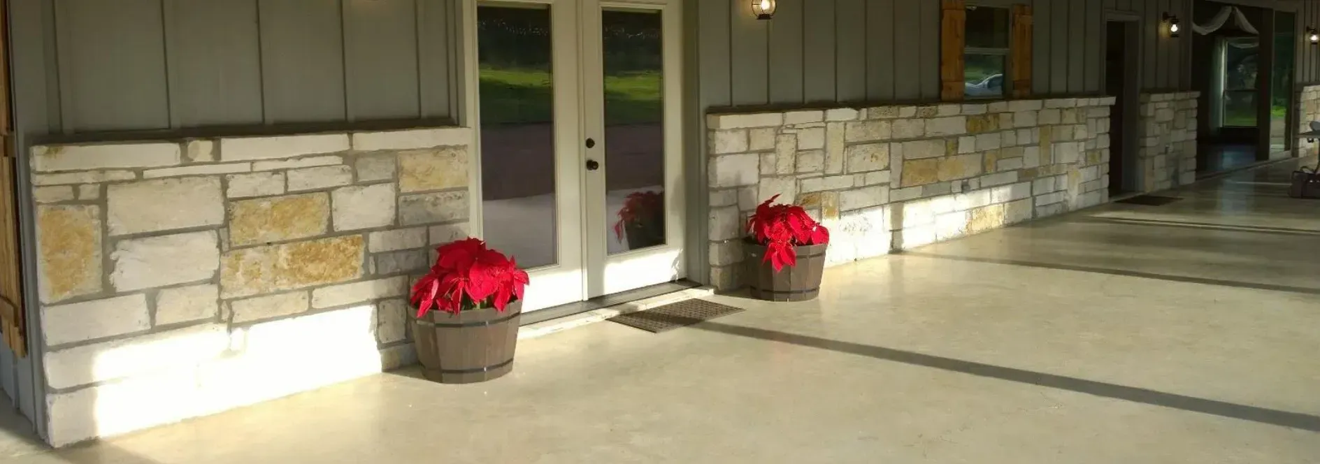 Front entrance with white double doors, stone half-wall, and two red flower pots in sunlight