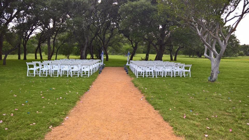 White chairs line a dirt aisle outdoors beneath trees, set up for an outdoor ceremony.