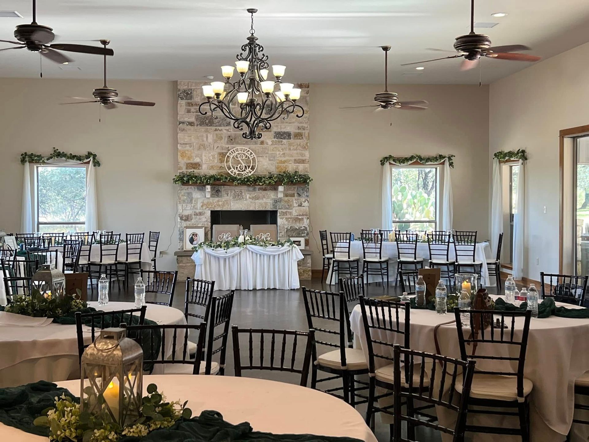 Empty banquet hall with chandeliers, ceiling fans, and round tables with chairs on a dark floor