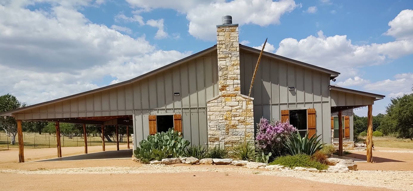 Rural metal house with stone chimney, porch, and flowering shrubs under a cloudy sky