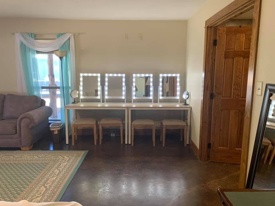 Dressing table with three lit mirrors against a beige wall, wooden door at right, dark floor.