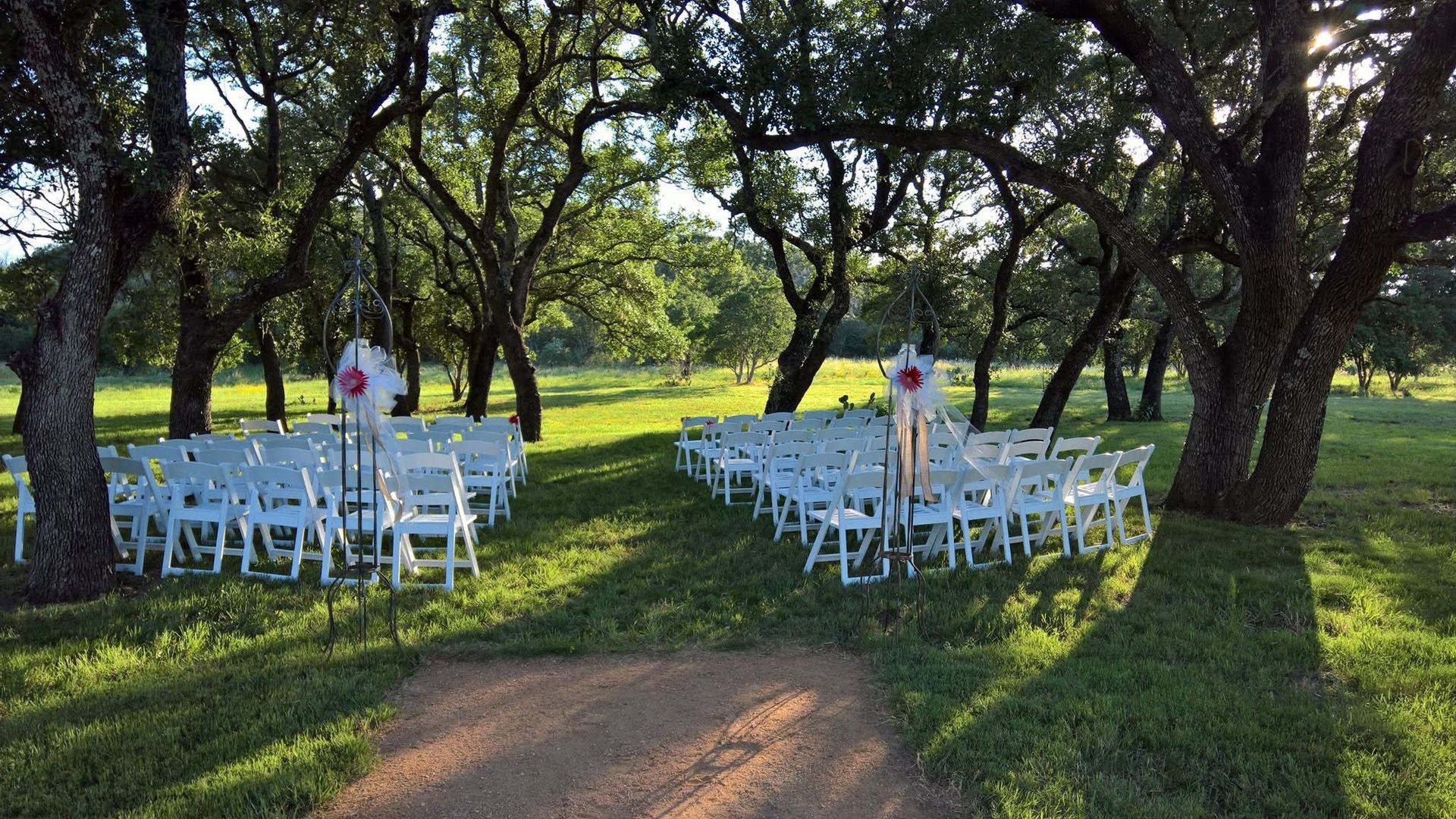 Outdoor wedding ceremony setup with white chairs under trees and a grassy aisle in sunlight