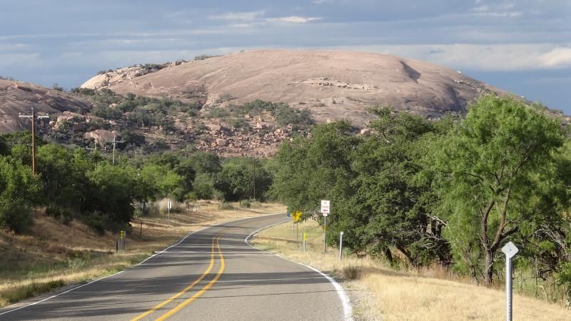 Winding road through green trees toward a large rocky hill under a partly cloudy sky
