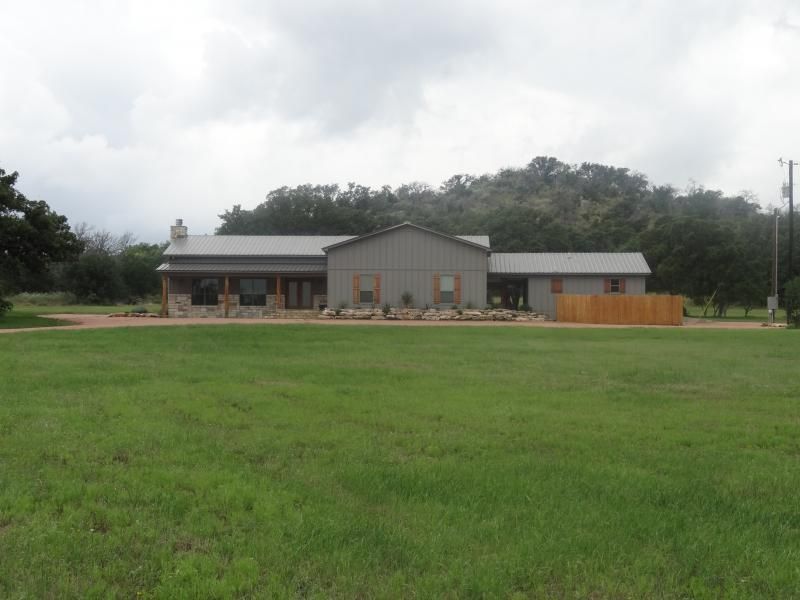 Gray building with a porch and wooden fence behind a large green field under a cloudy sky