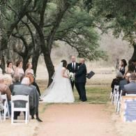 Outdoor wedding ceremony with bride and groom under trees, guests seated on both sides.