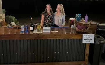 Two women stand behind a vendor table with snacks and drinks in a dimly lit event space.