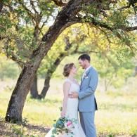 Bride and groom standing under a tree in a sunlit outdoor setting