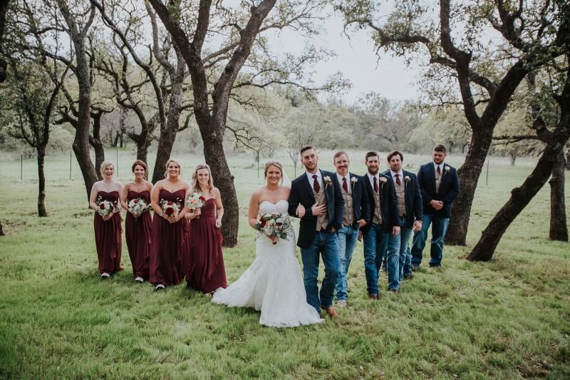 Wedding party posing in a grassy field among trees, with bride in white and attendants in burgundy and navy suits