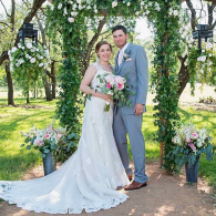 Bride and groom pose in a flowered garden arch, holding a bouquet on their wedding day.