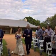 Bride and groom walk down an outdoor aisle with guests seated on white chairs under a cloudy sky.