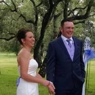 Bride and groom standing outdoors under trees, smiling and holding hands at a wedding ceremony