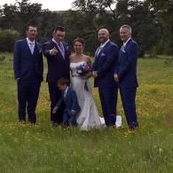 Wedding party posing outdoors in a grassy field, with bride, groom, groomsmen, and a child in front.
