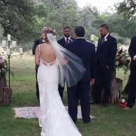 Bride in white dress and veil standing at outdoor wedding ceremony with groom and officiant